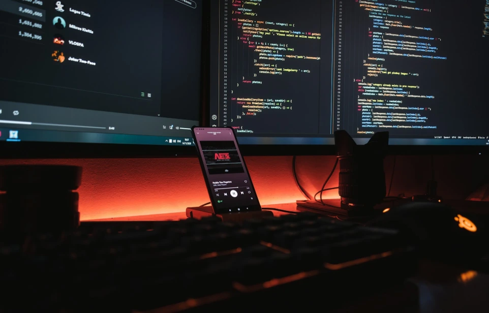A smartphone displaying music on a desk with computer monitors showing code